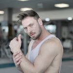 Side view of strong male athlete in casual sportswear standing with raised crossed hands near chest while resting after workout in modern fitness center and looking at camera
